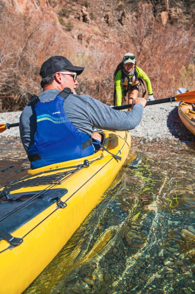 Man in blue vest paddles yellow kayak on clear rocky water, woman assists from shore.