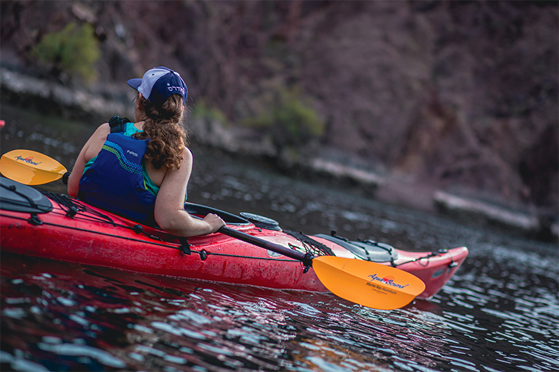 Person in a red kayak on a calm lake near rocky cliffs, wearing a blue life jacket and cap.
