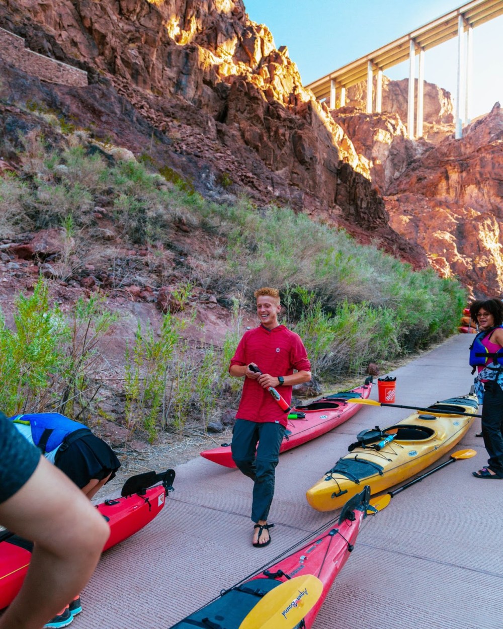 People near colorful kayaks on a riverside path, with a rocky cliff and bridge in the background.