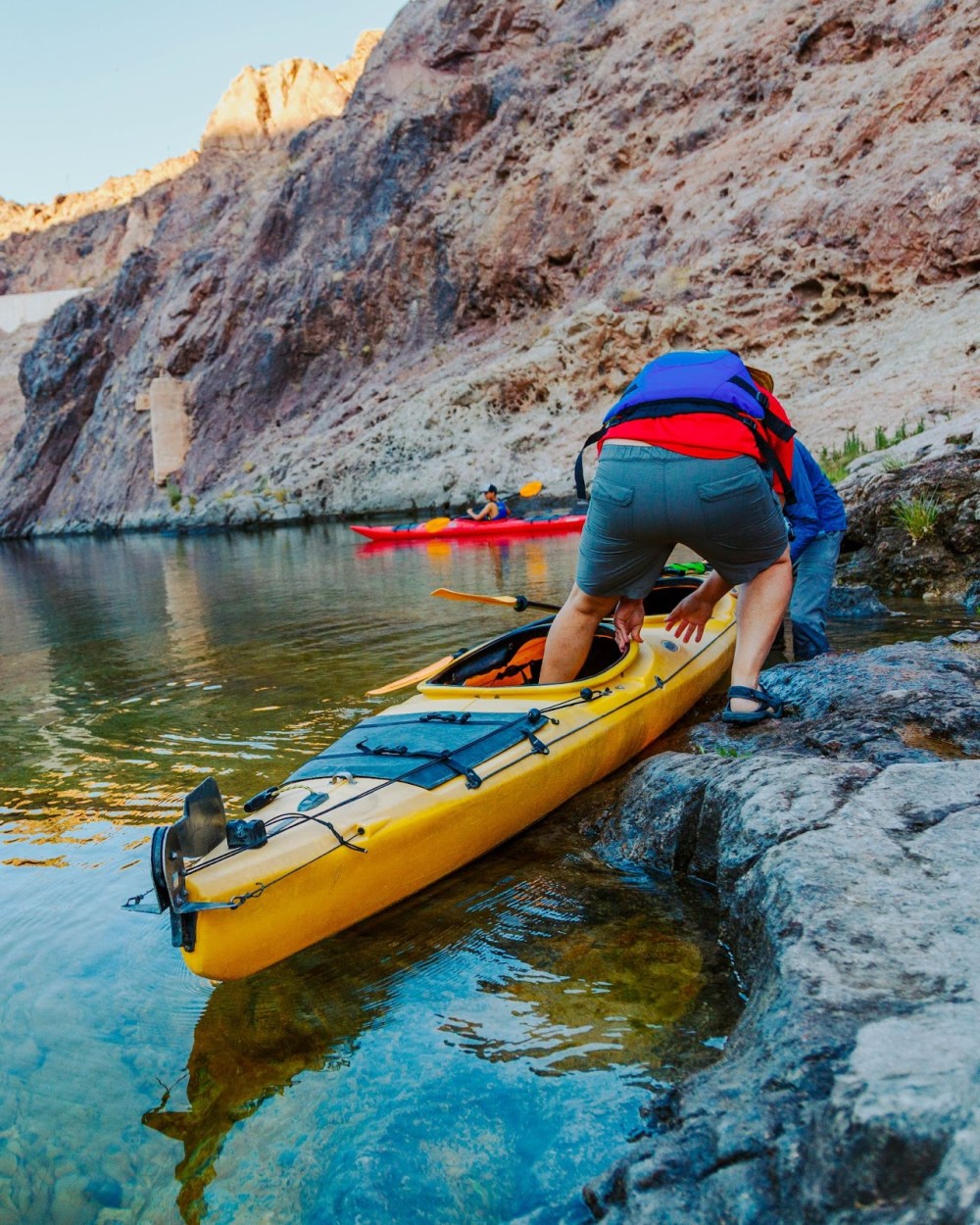 Person launching a yellow kayak on rocky shore beside cliffs, with others kayaking in the background.