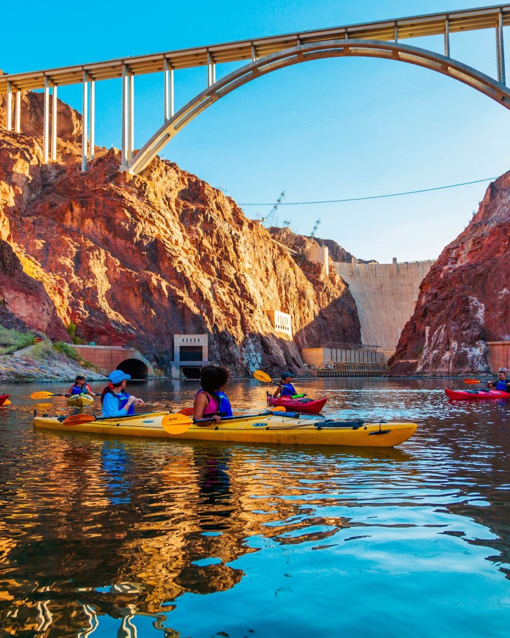 Kayakers on a river beneath a large, arched bridge near red rocky cliffs.