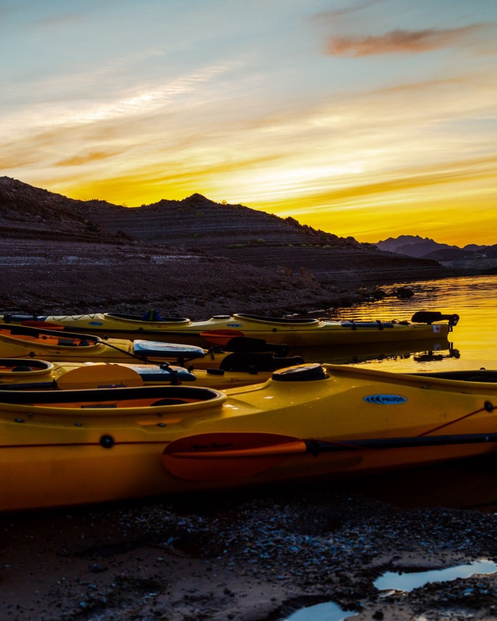 Yellow kayaks lined up on rocky shore at sunset with mountains in background.