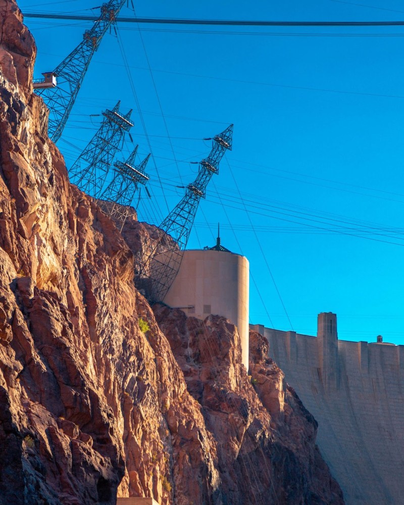 Rocky cliffs with power lines and a dam structure under a clear blue sky.