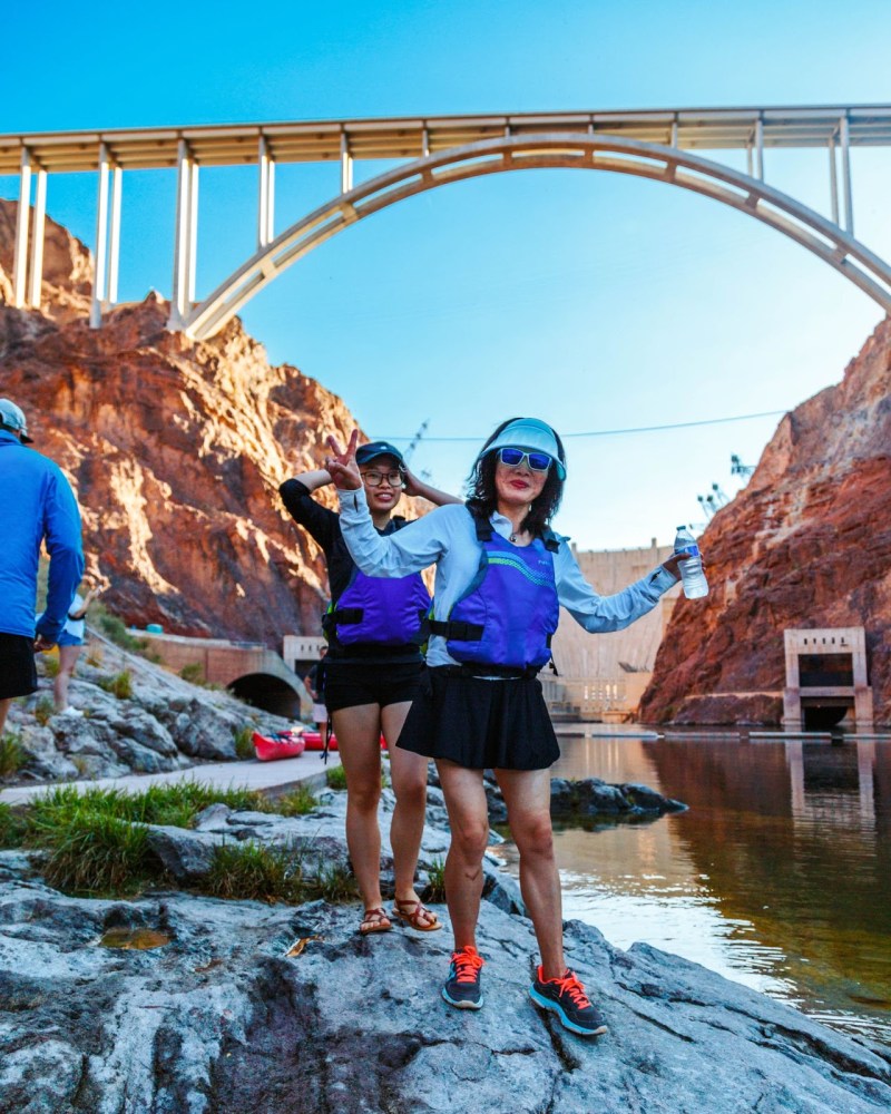 Two women in life jackets posing by a river under a large bridge on a sunny day.