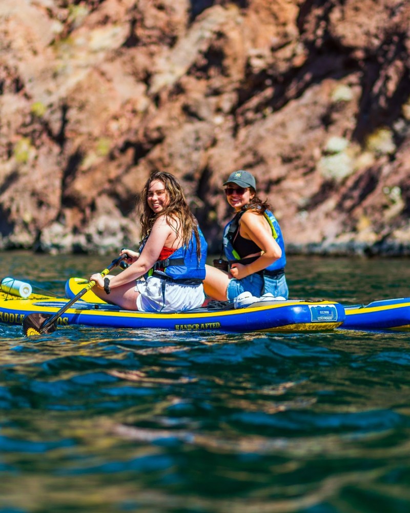 Two women in life vests kayaking on a blue inflatable kayak near rocky cliffs.
