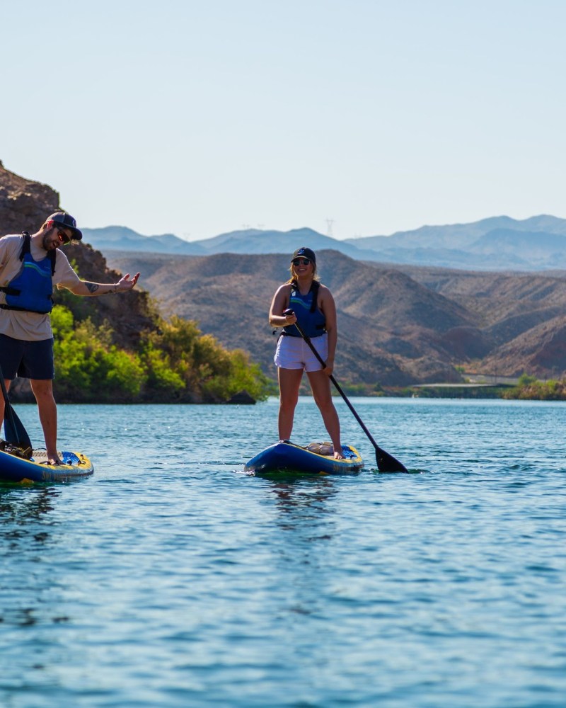 Two people paddleboarding on a calm river with mountainous backdrop.