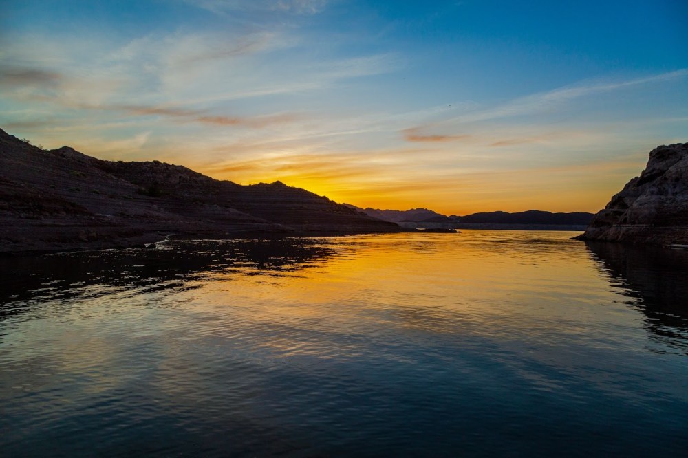 Sunset over a calm lake with silhouetted hills and a colorful sky.
