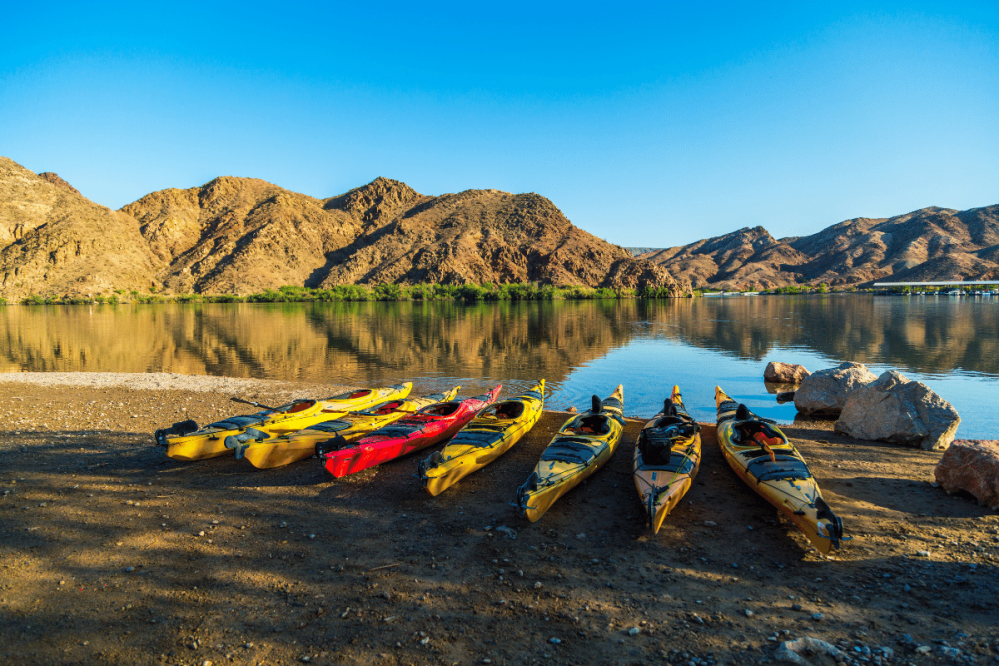 Kayaks on a beach by a calm lake with hills in the background under a clear blue sky.
