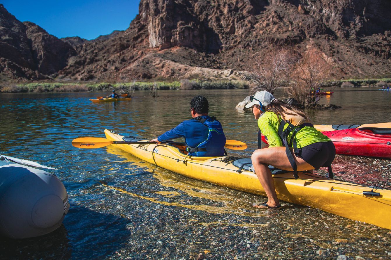 Two people in a yellow kayak on a clear river with rocky banks and a mountain backdrop.