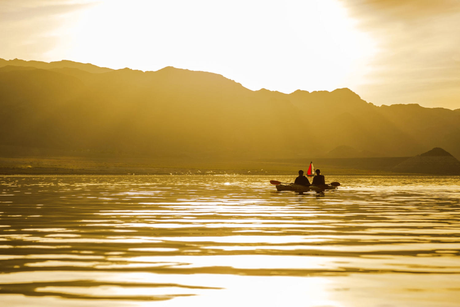 Two people in a kayak on a lake at sunset with mountain silhouette in the background.