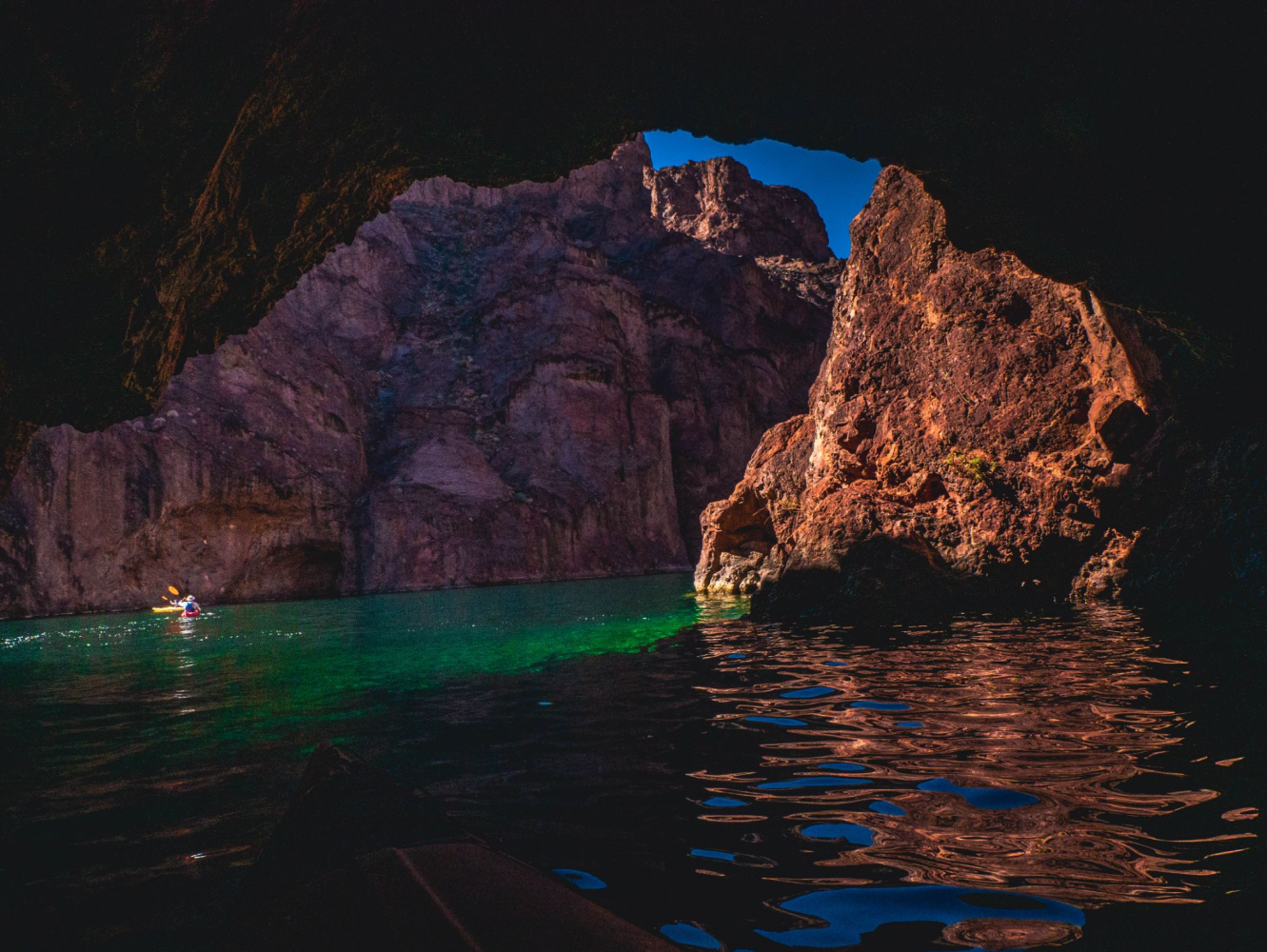 Cave entrance with sunlit rock formations and a person kayaking on a greenish-blue water surface.
