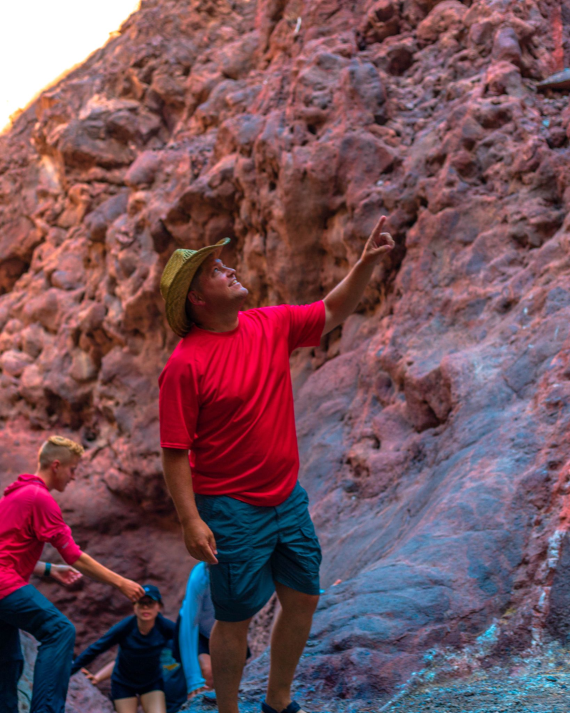Man in red shirt and hat pointing at rocky cliffs, with climbers in background.