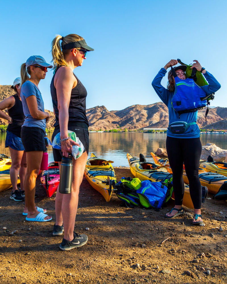 Four people near kayaks preparing by a calm lake with mountains and clear blue sky.