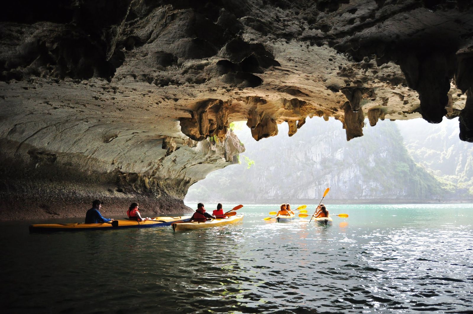 Kayakers paddle through a cave with stalactites, entering a sunlit lagoon.