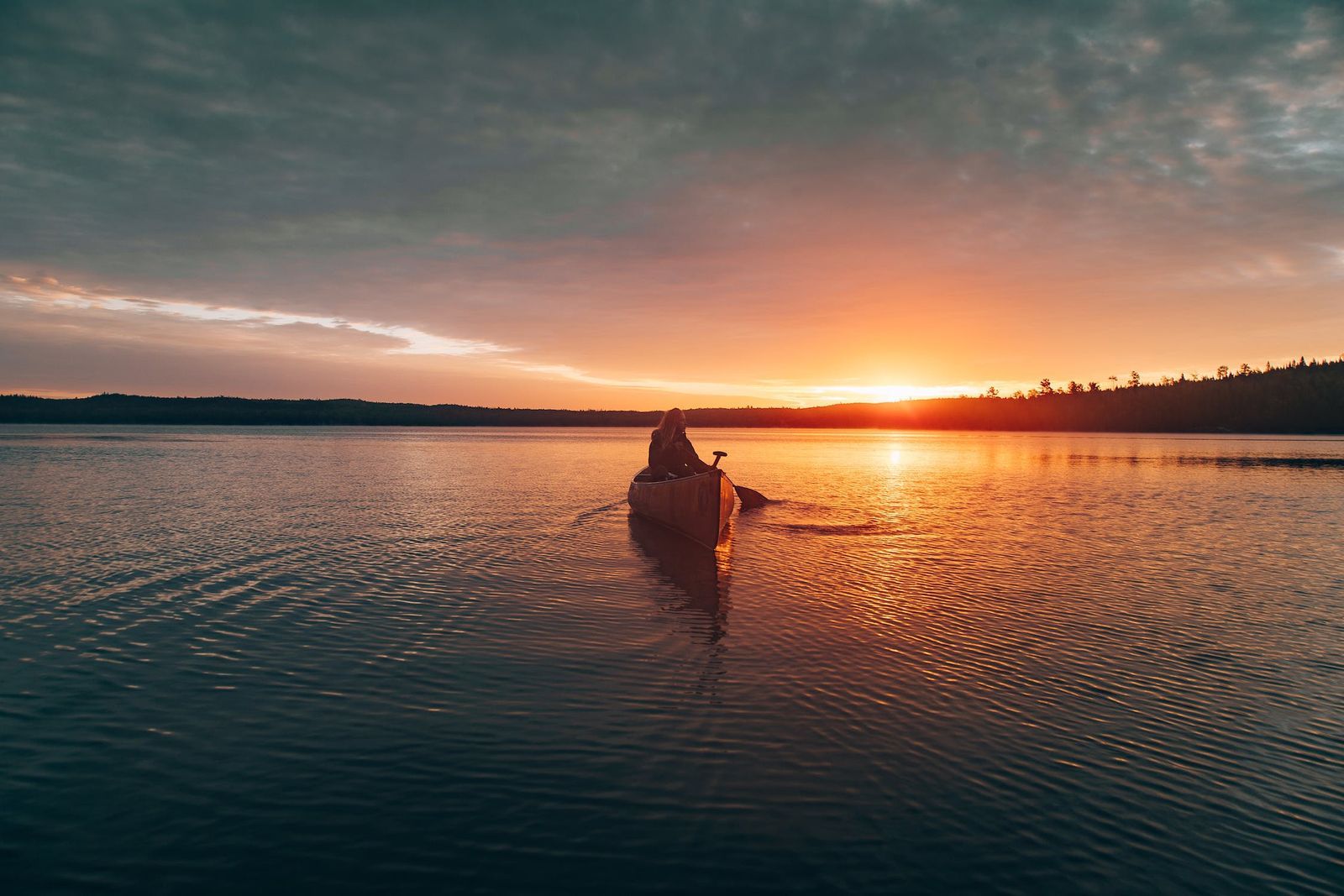 Person canoeing on a lake during sunrise or sunset with cloudy sky.