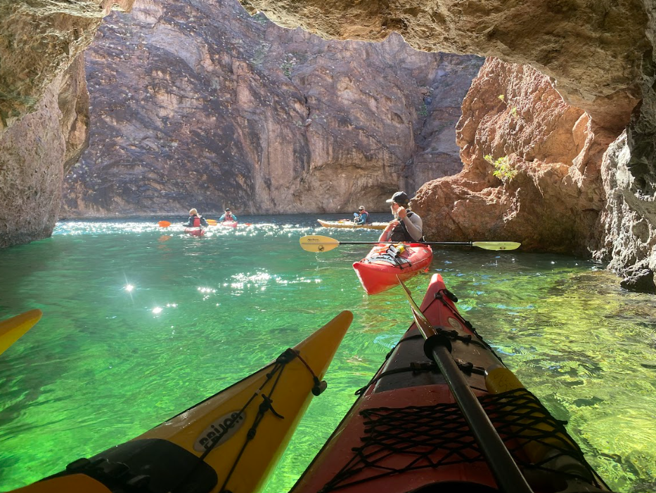 People kayaking on clear green water inside a rocky cave with sunlight.