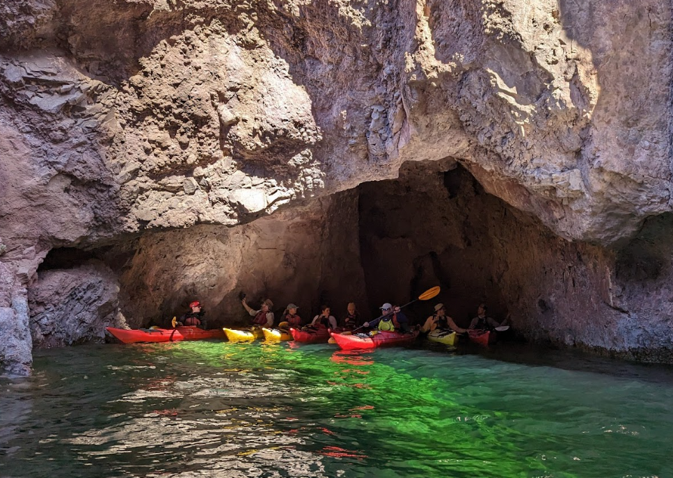 Group kayaking into a cave with rocky walls and sunlight illuminating green water.