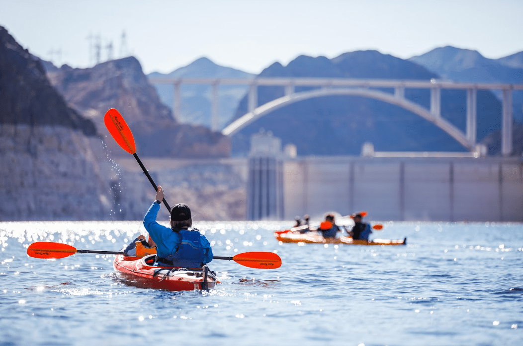 Kayakers paddling on a lake with a large bridge and rocky landscape in the background.