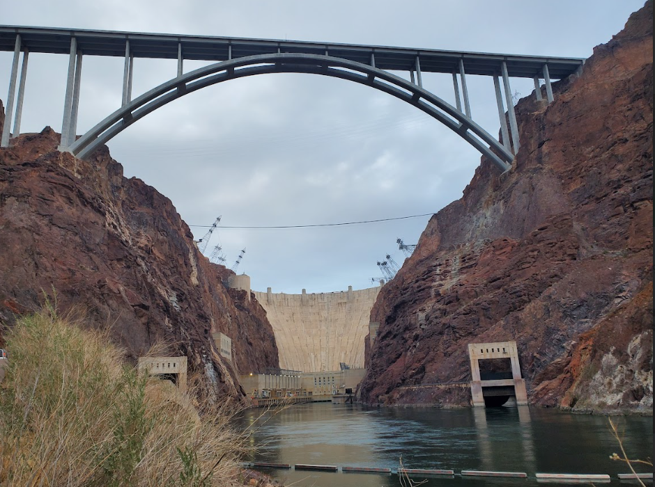 Arched bridge over rocky canyon with dam and water below on a cloudy day.