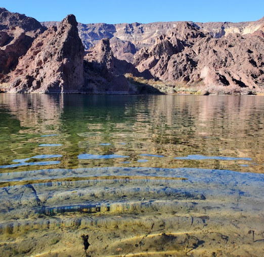 Clear water lake with ripples and rocky mountain backdrop under blue sky.
