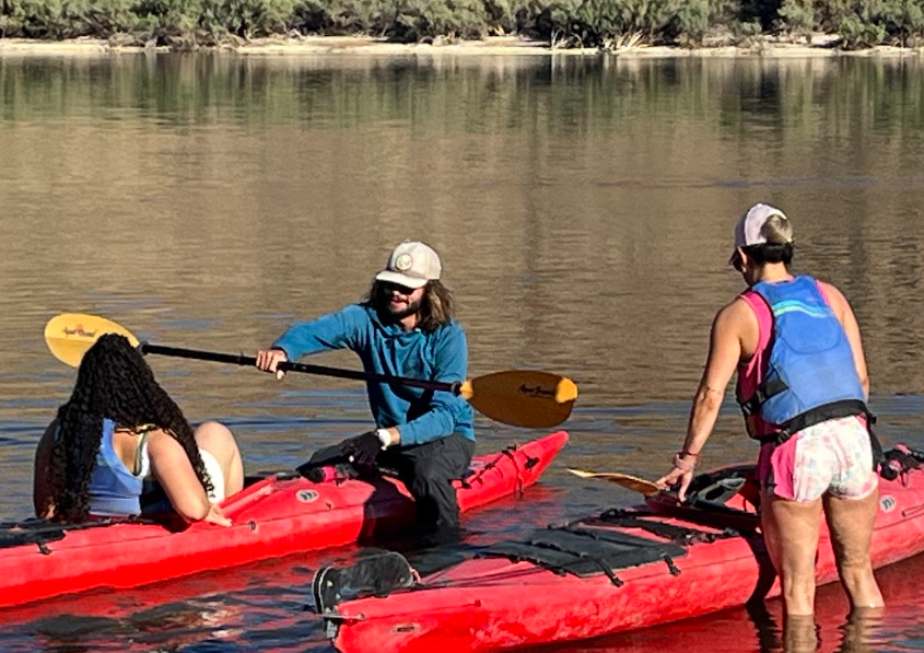 Three people on red kayaks in a calm river, with trees in the background.