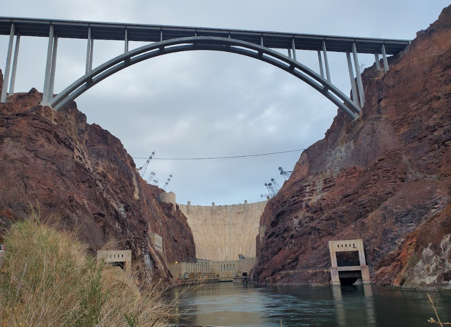 View of a large dam between rocky cliffs with an arch bridge above.