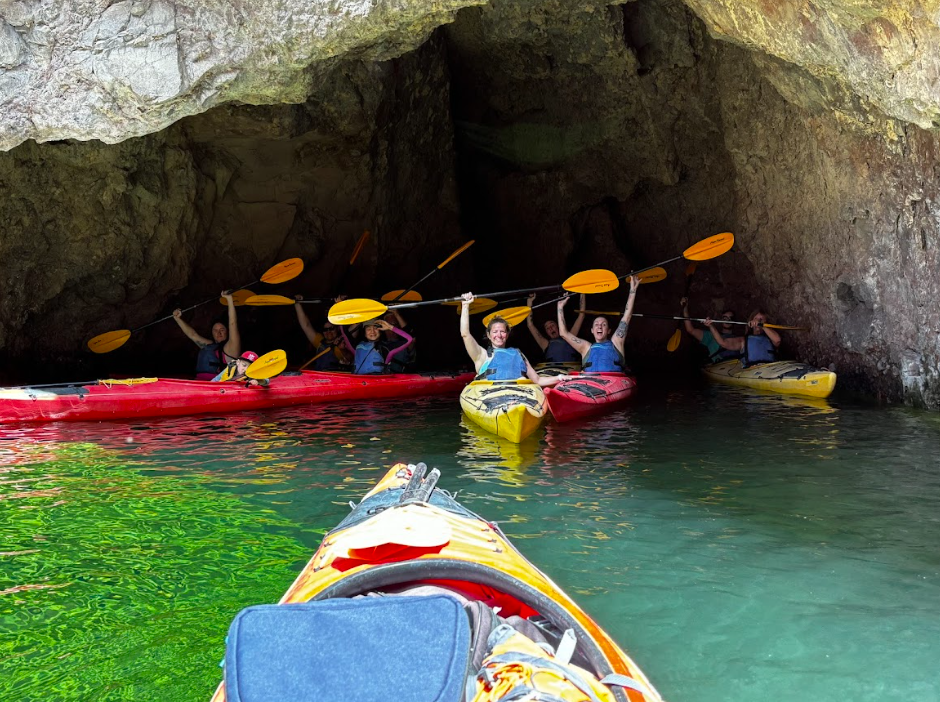 People in kayaks raising paddles inside a rocky cave.
