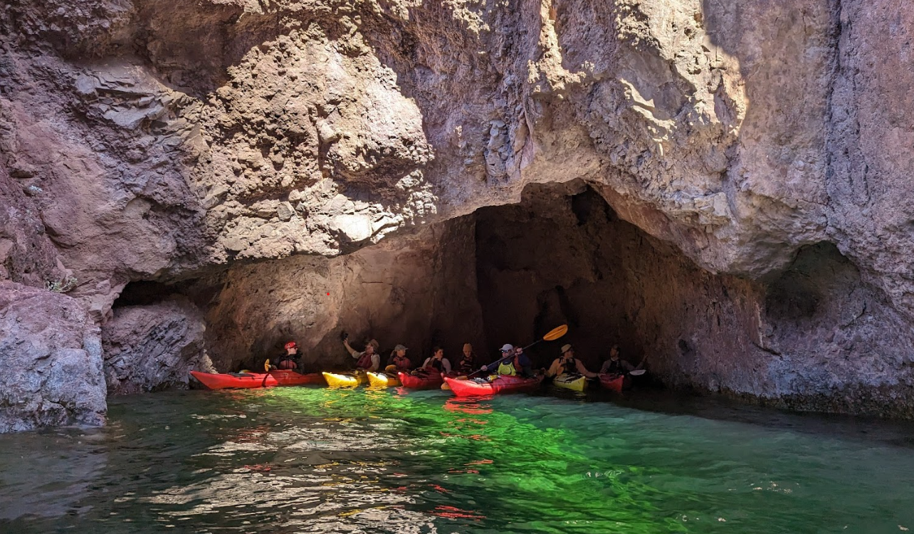 Kayakers in colorful kayaks near a rocky cave entrance on a sunny day.