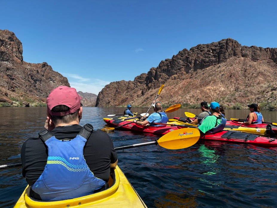 Group kayaking on a calm river with rocky cliffs under a clear blue sky.