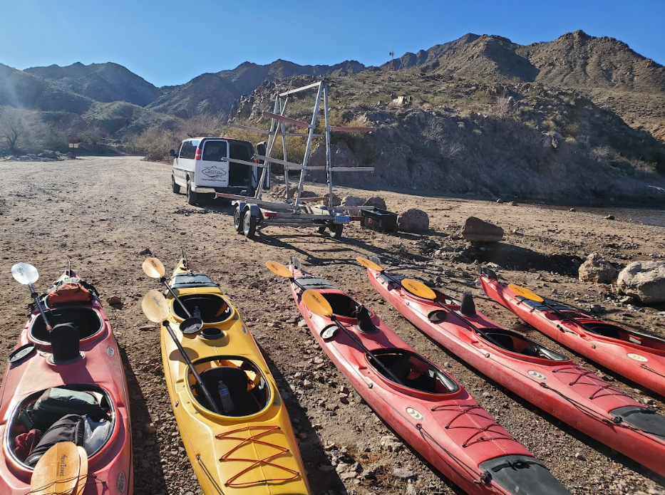Kayaks on rocky shore with van and mountains in background.