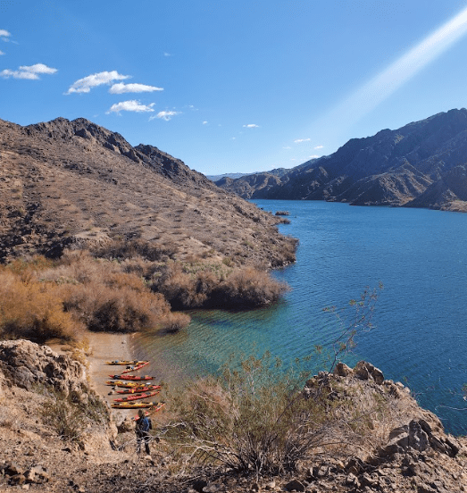 Kayaks on a small beach by a blue lake surrounded by rocky hills under a clear sky.