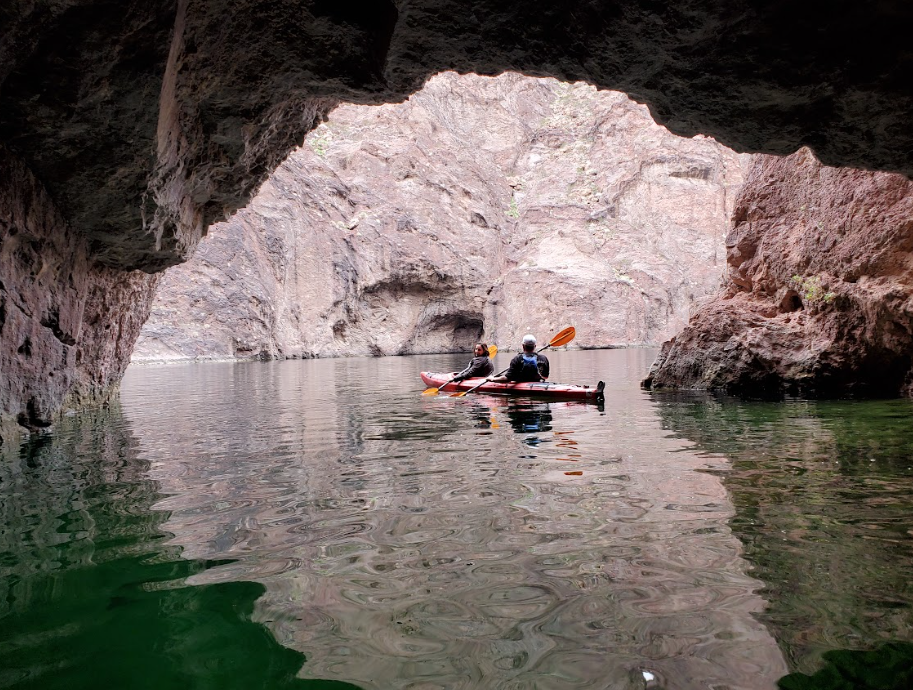 Two people kayaking in a cave with rocky walls and green water.