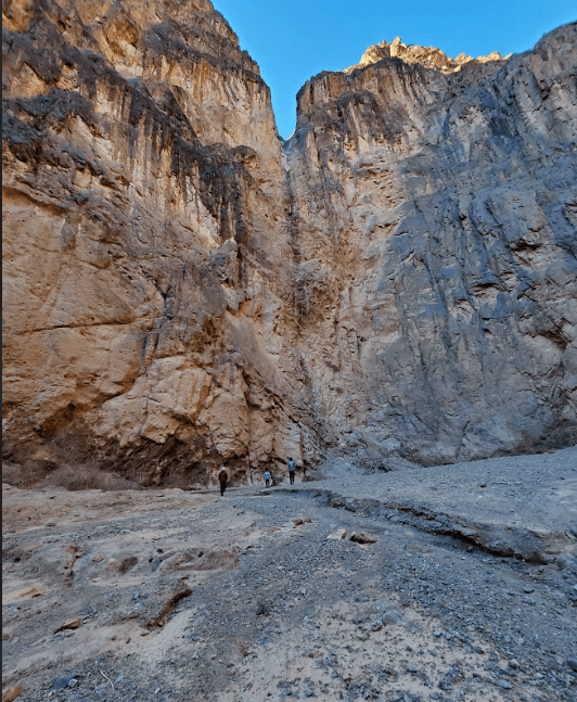 People hiking in a narrow canyon with steep rocky walls under a clear blue sky.
