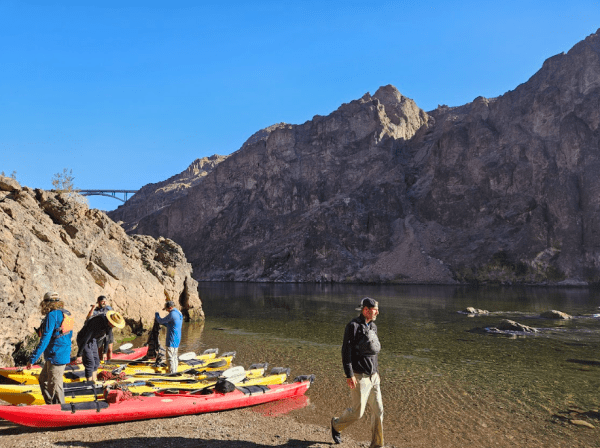 People with kayaks on a rocky shore near a river, with a bridge and mountains in the background.