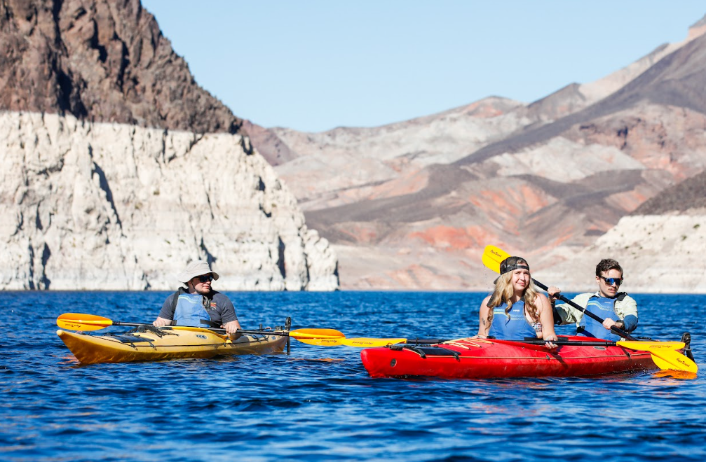 Two kayaks on a lake with mountainous backdrop; three people paddling.