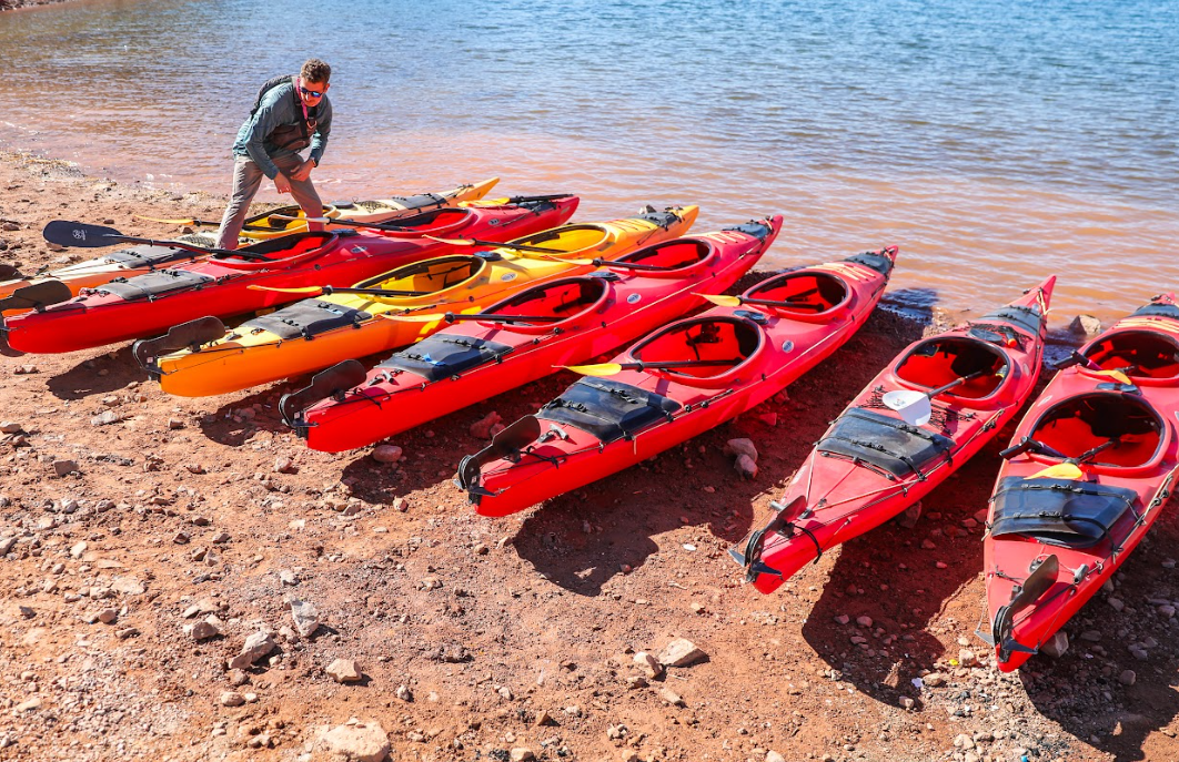 Person organizing red and yellow kayaks on a rocky lakeshore.