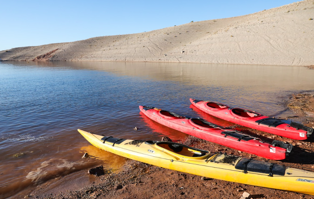 Three kayaks, two red and one yellow, on a sandy shore by a calm lake under a clear sky.