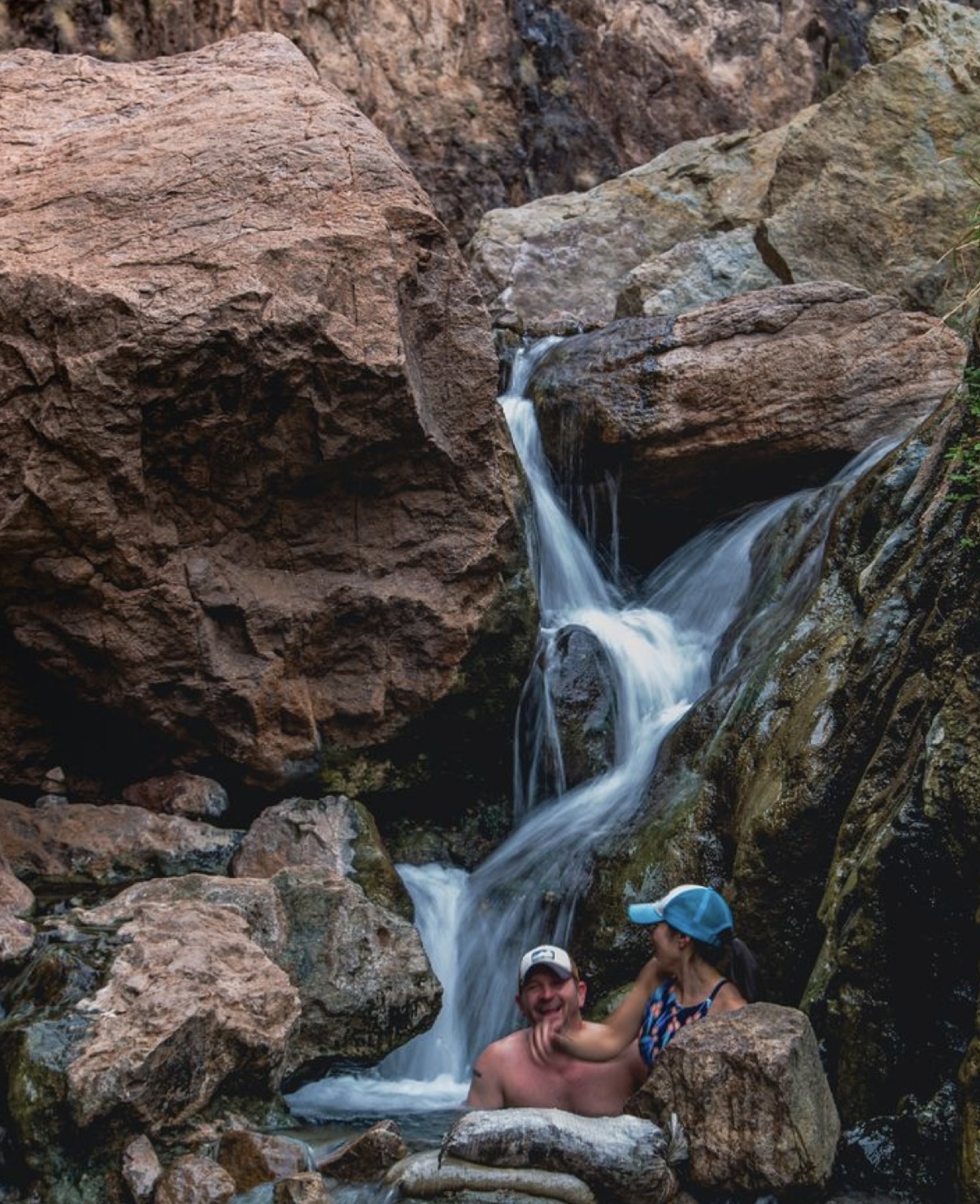 Two people relaxing in a small waterfall surrounded by large rocks.