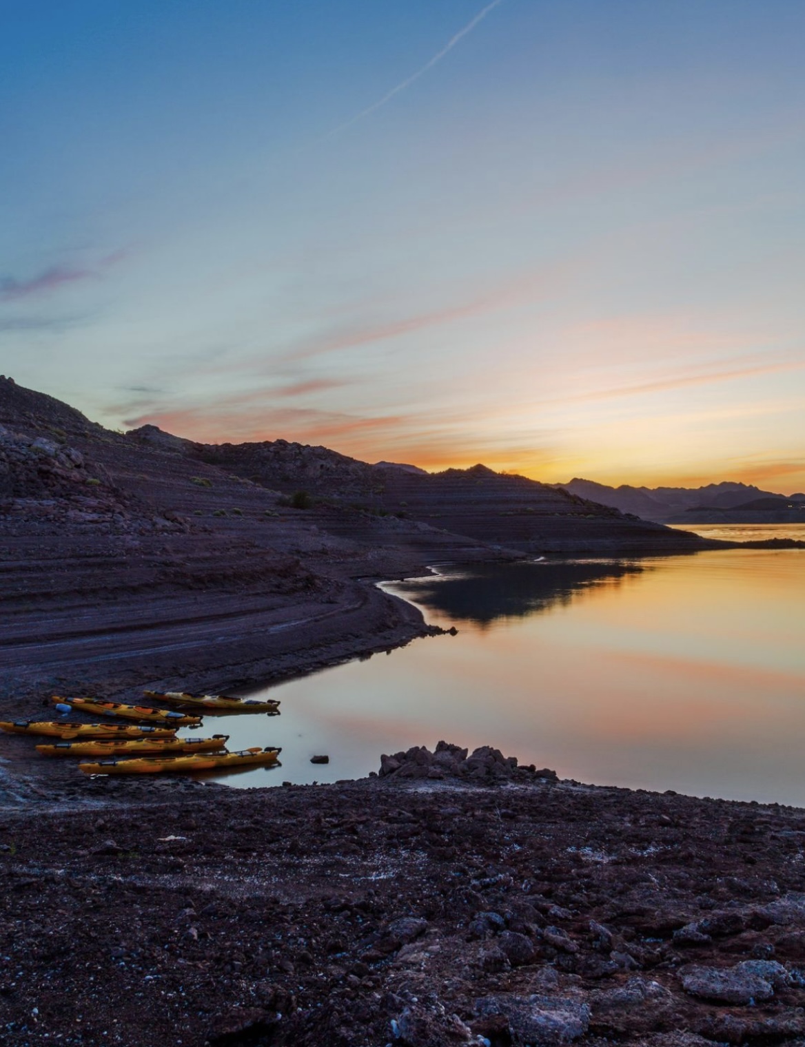 Sunset over a rocky shore with yellow kayaks and calm water reflecting colorful sky.