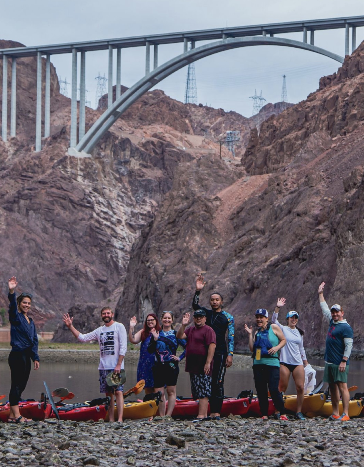 Group of people waving next to kayaks by a river under a large bridge.