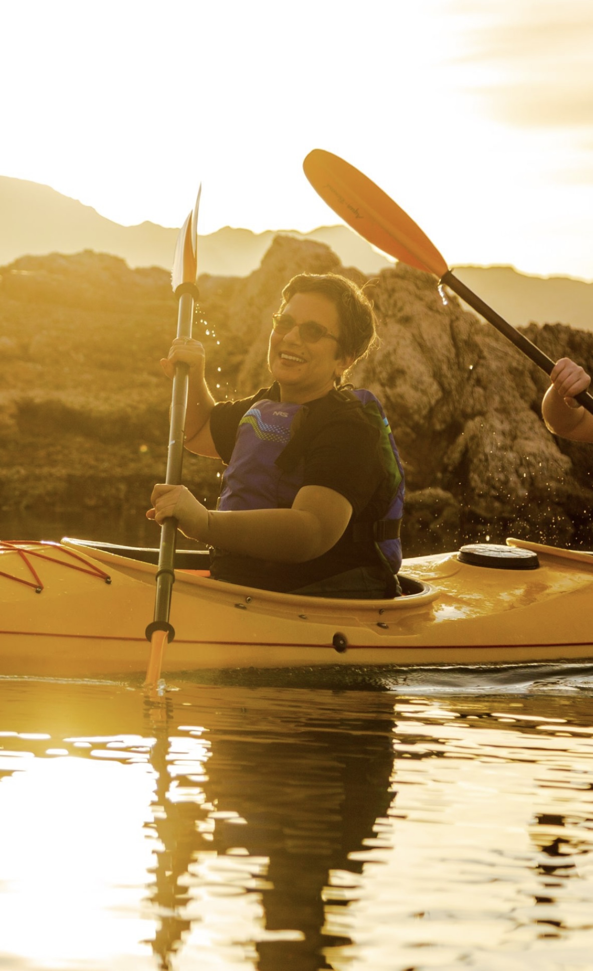 Person kayaking on calm water at sunset, smiling and wearing sunglasses.