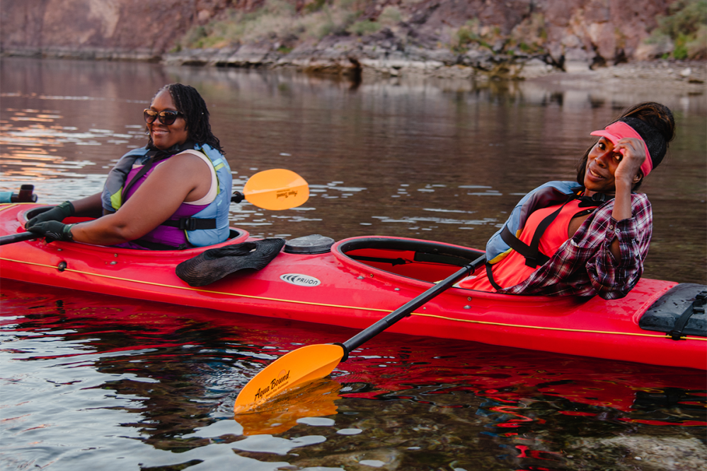 Two people kayaking on a calm river in red kayaks, wearing life vests and casual clothing.