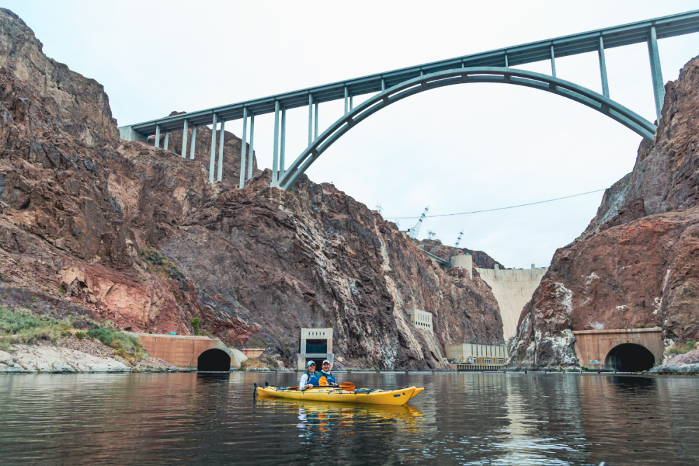 Two people kayaking under a bridge near rocky cliffs and a dam.