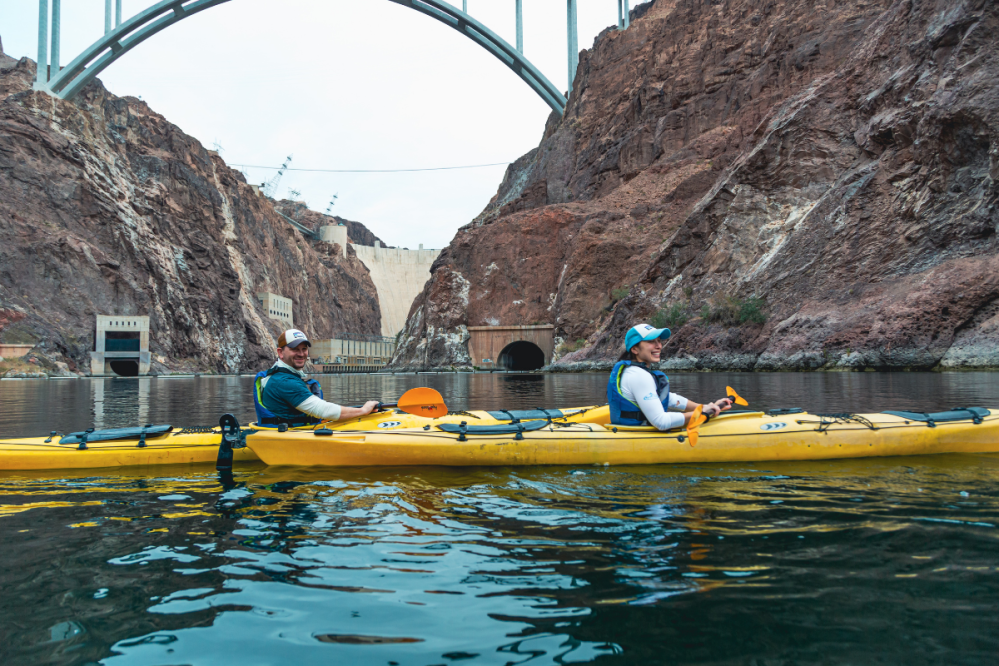 Two people kayaking on a river with a large dam and bridge in the background.