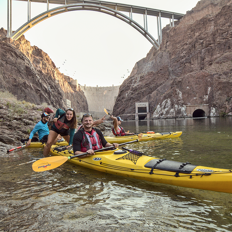 People kayaking on a river beneath a large arched bridge and rocky cliffs.