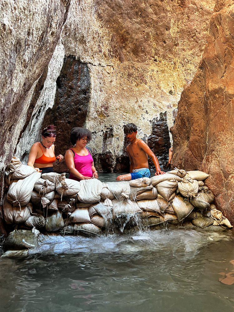 Three people sitting on sandbags by a rocky pool with water cascading over the bags.