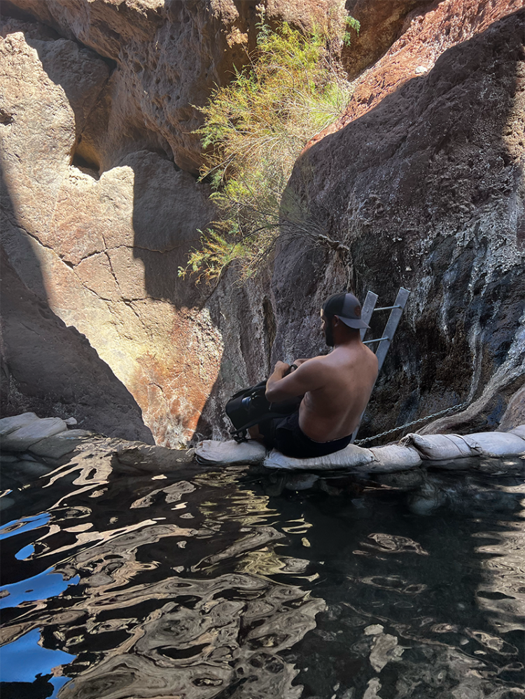 Man sitting on rocks by water in a rocky canyon, wearing a cap.
