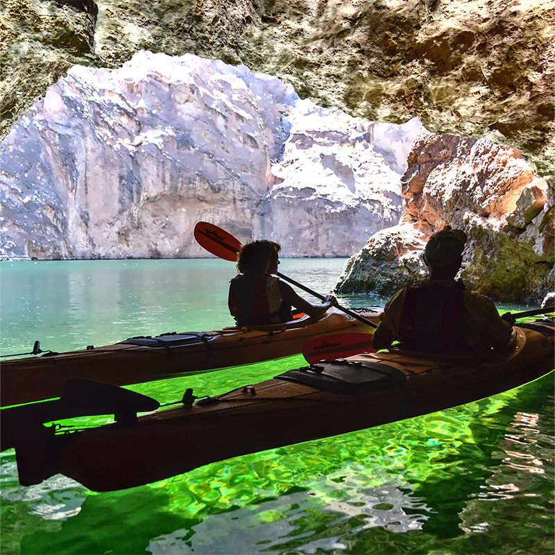 Two people kayaking in a cave with sunlit water and rocky walls.