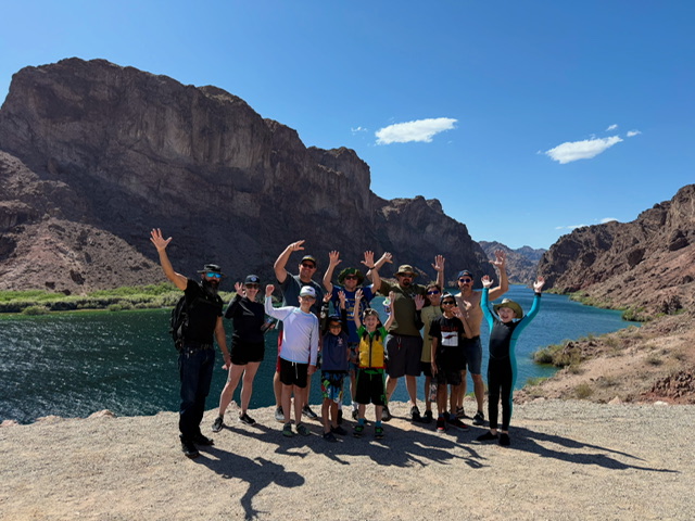 Group of people waving by a river with rocky cliffs under a clear blue sky.