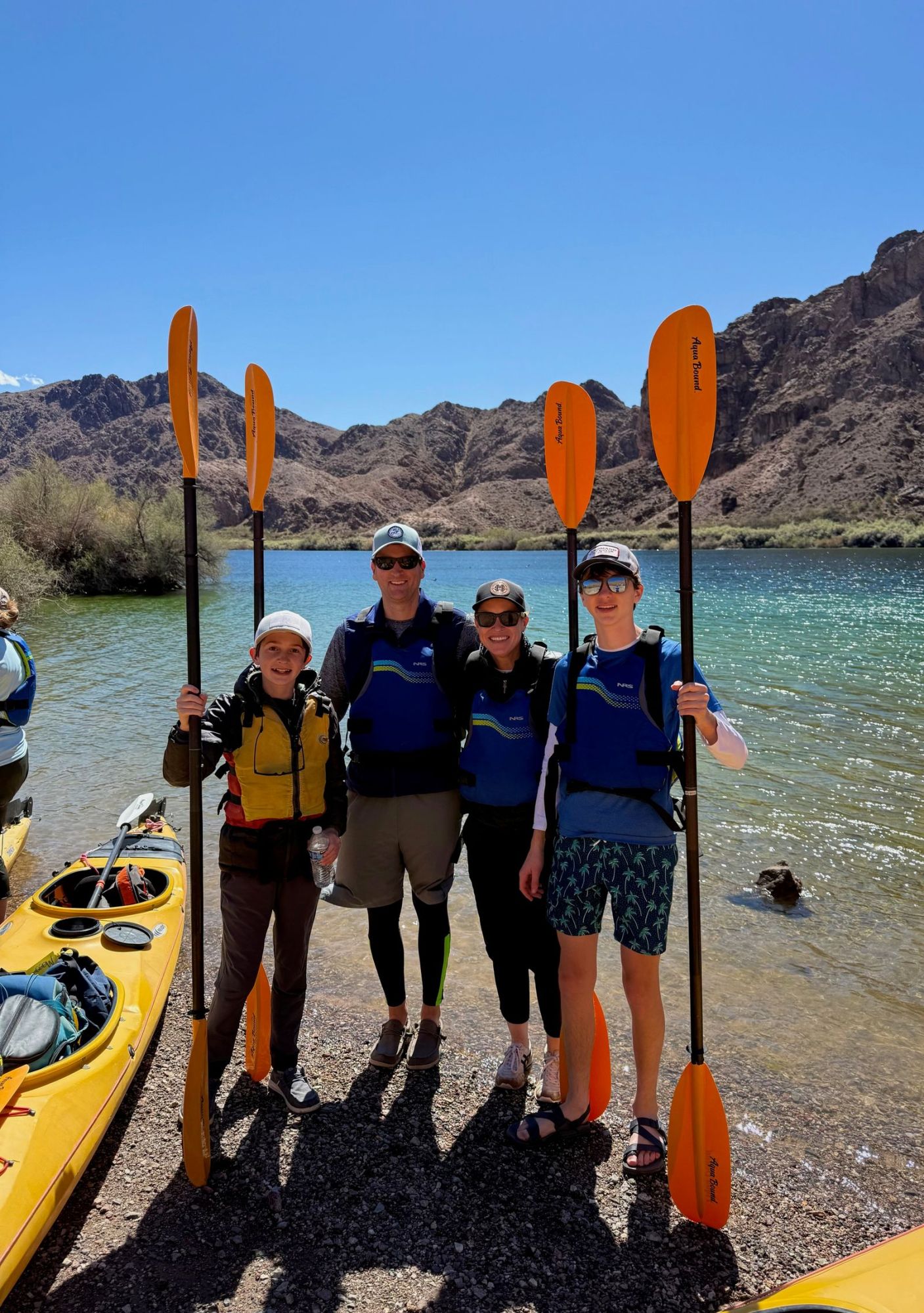 Four people in life vests hold kayak paddles by a lake with mountains in the background.